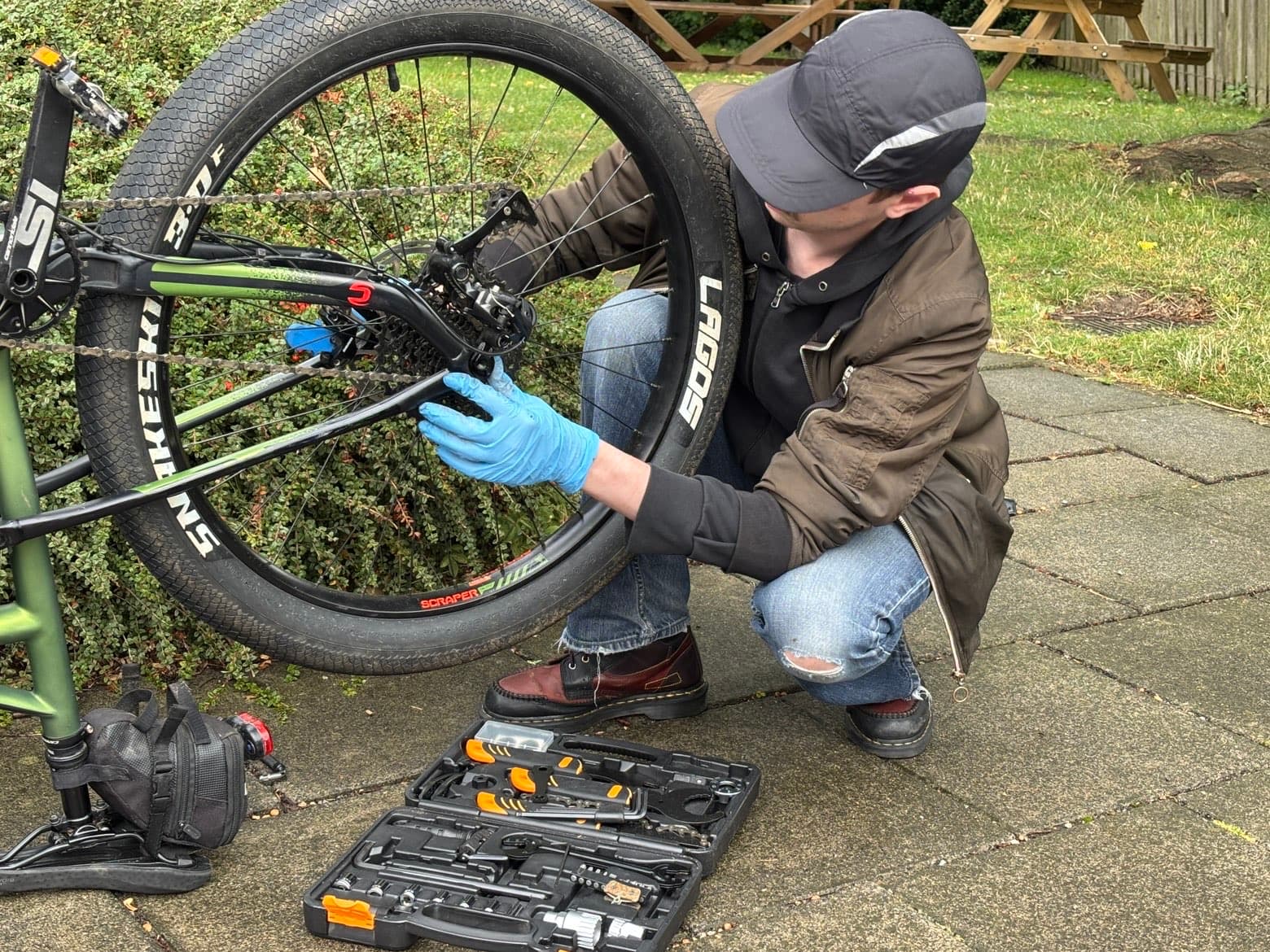 Mechanic adjusting a bicycle rear derailleur outdoors, with a tool case beside the bike